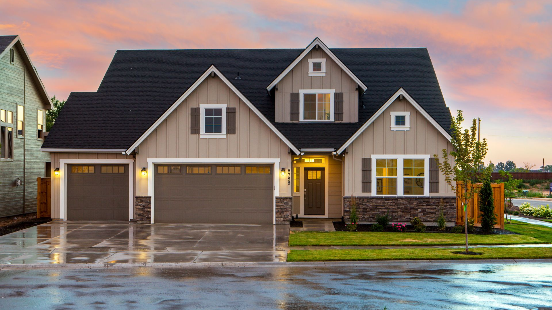 Suburban house with three garage doors, lit porch, and wet driveway at sunset