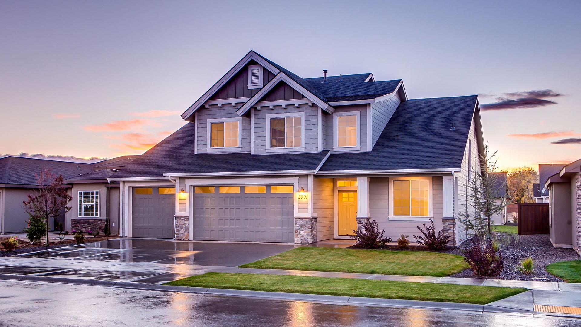 Suburban house at dusk with lit windows, wet driveway, and a reflective street after rain