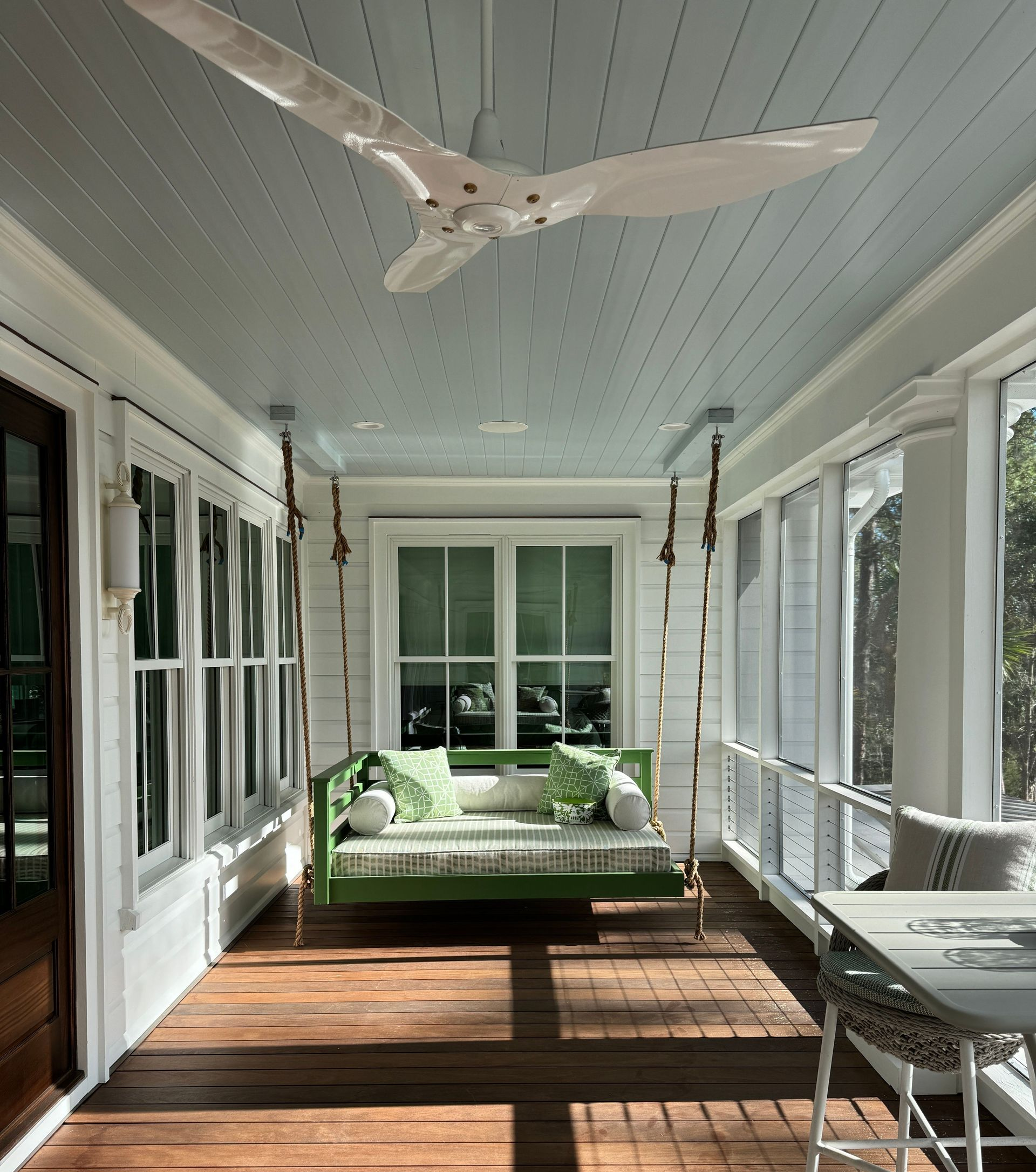 Covered porch with white ceiling fan, wooden floor, wicker furniture, and a green daybed in bright sunlight