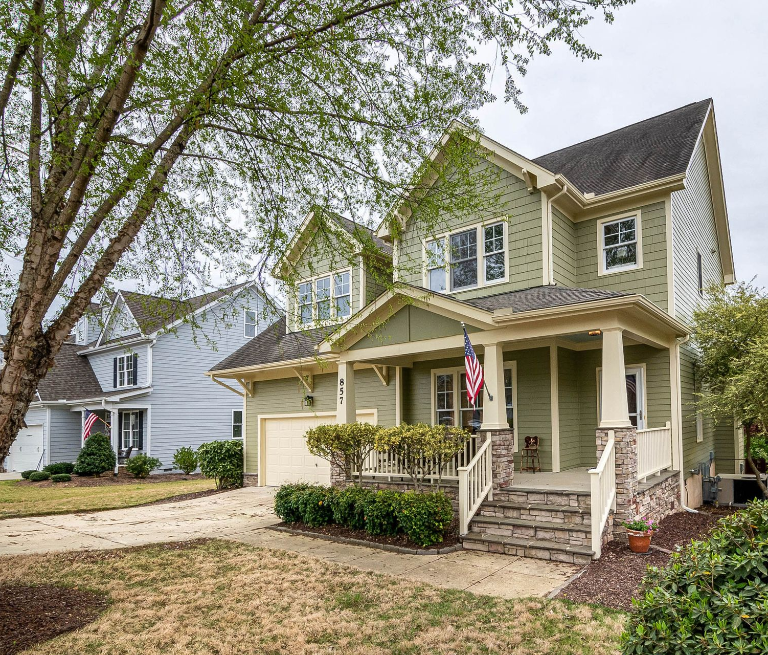 Yellow two-story house with a front porch, American flag, and driveway surrounded by trees and shrubs