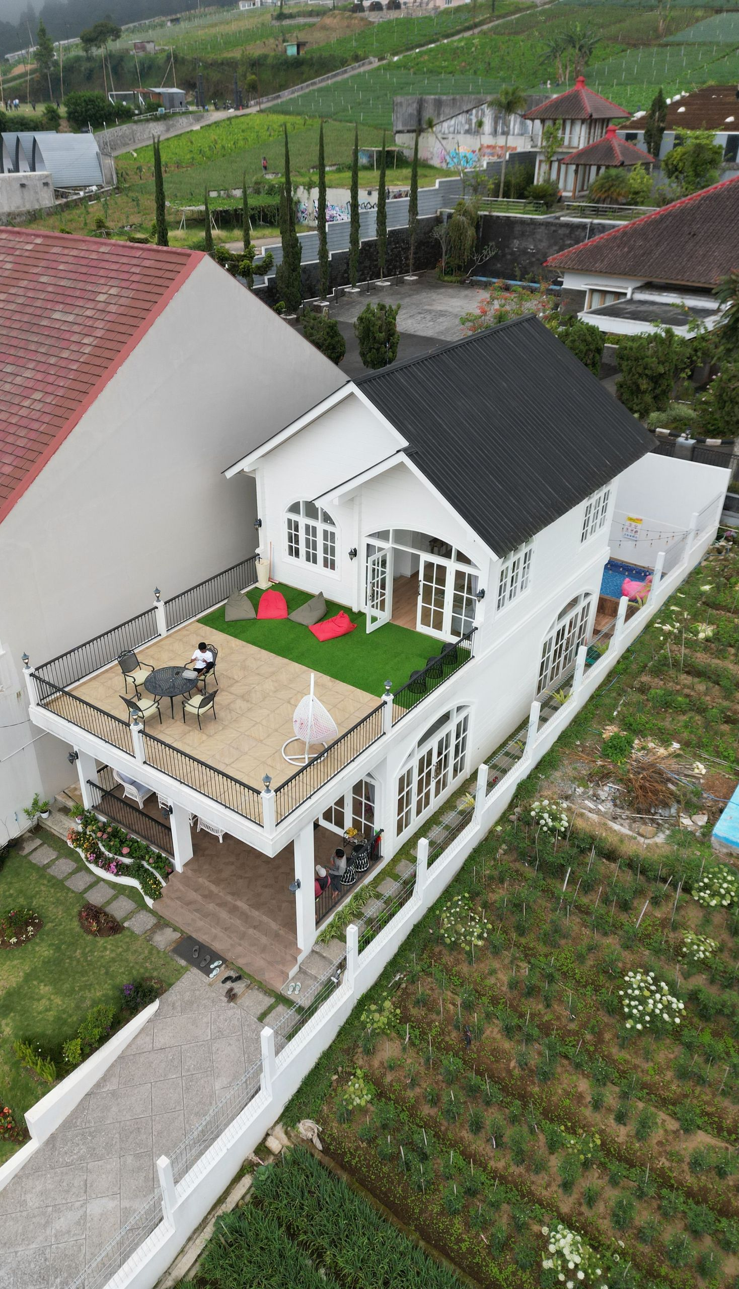 Aerial view of a white house with a rooftop terrace beside green farm fields and neighboring homes