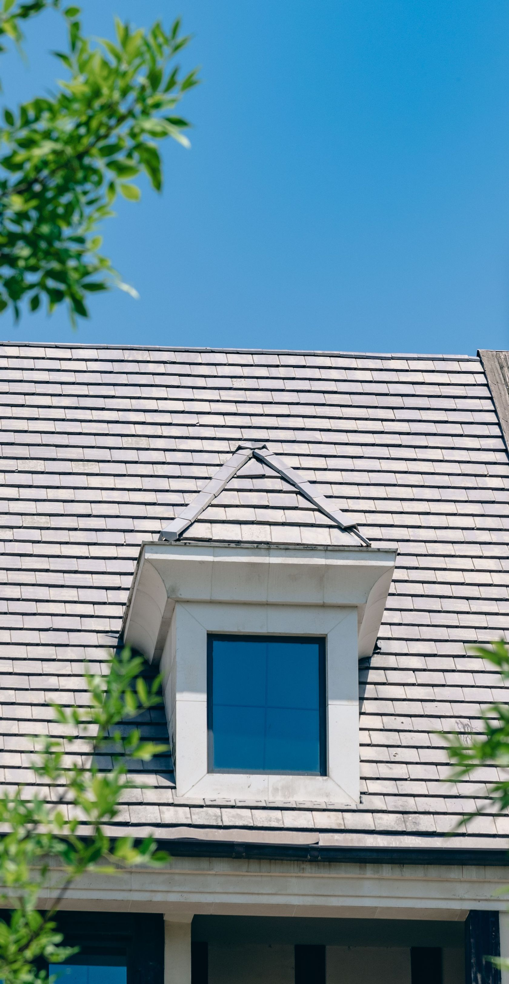 White house dormer with blue window under a clear blue sky, framed by green leaves