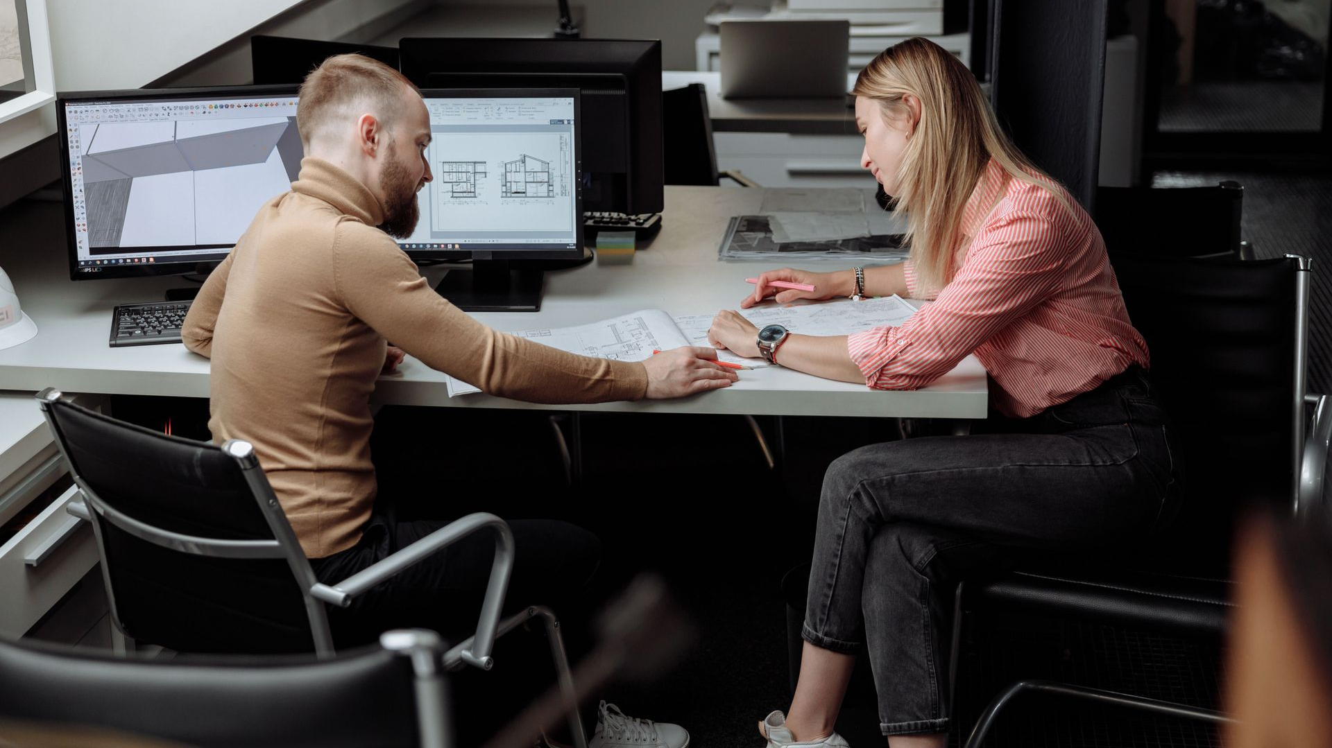 Two people collaborate at a desk with a laptop and monitors in a modern office
