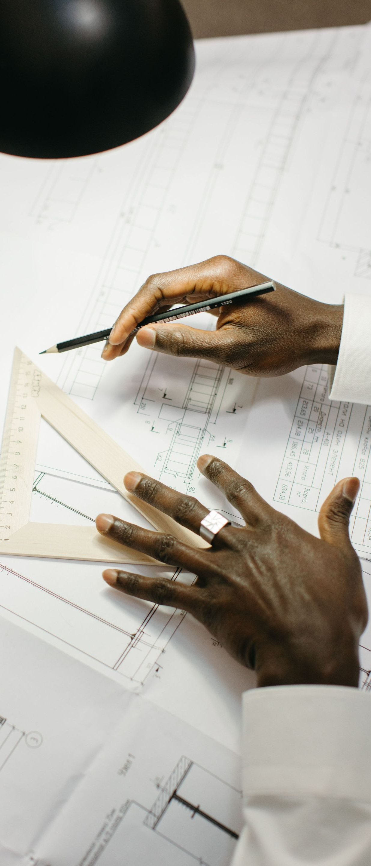 Hands sketching a design on white paper with a black pen under a desk lamp.