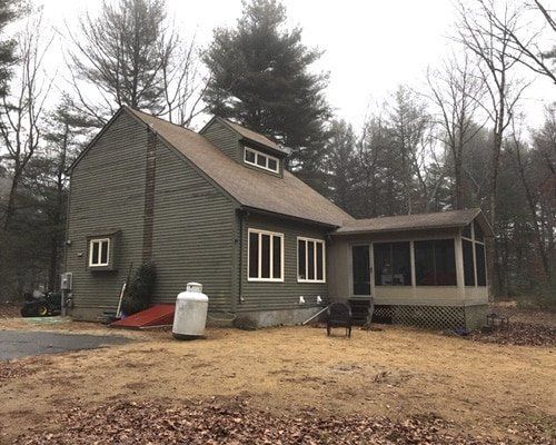 Gray house with steep roof and screened porch in a wooded yard on a cloudy day