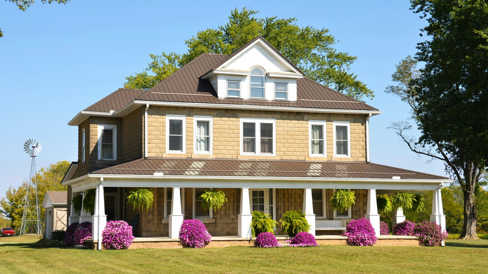 Two-story beige house with white porch, dormer windows, and pink flowers in front yard under blue sky