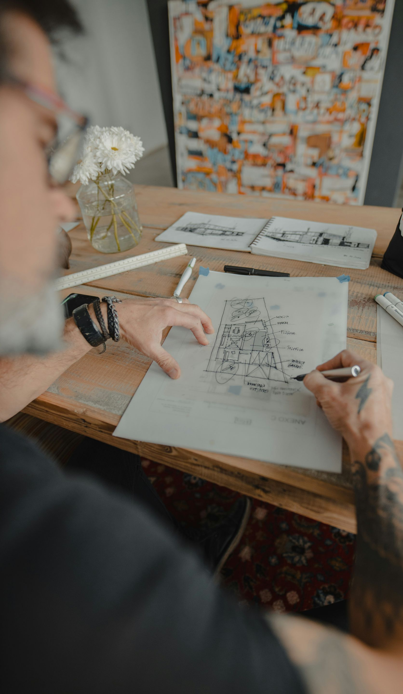 Two people sketching at a wooden table with flowers, papers, and art on the wall nearby