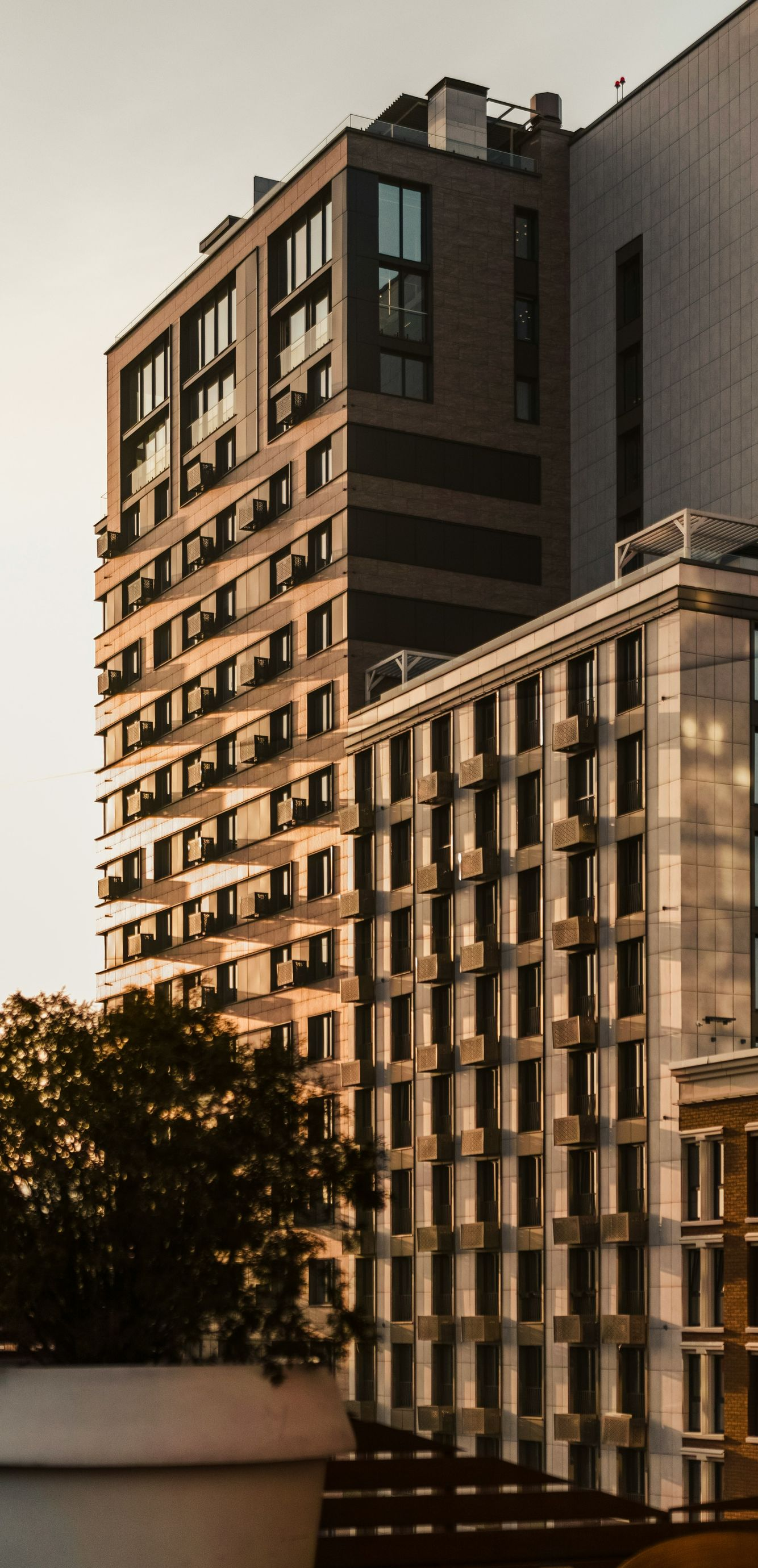 Urban apartment building with balconies at sunset, seen beside a tree and a white railing.