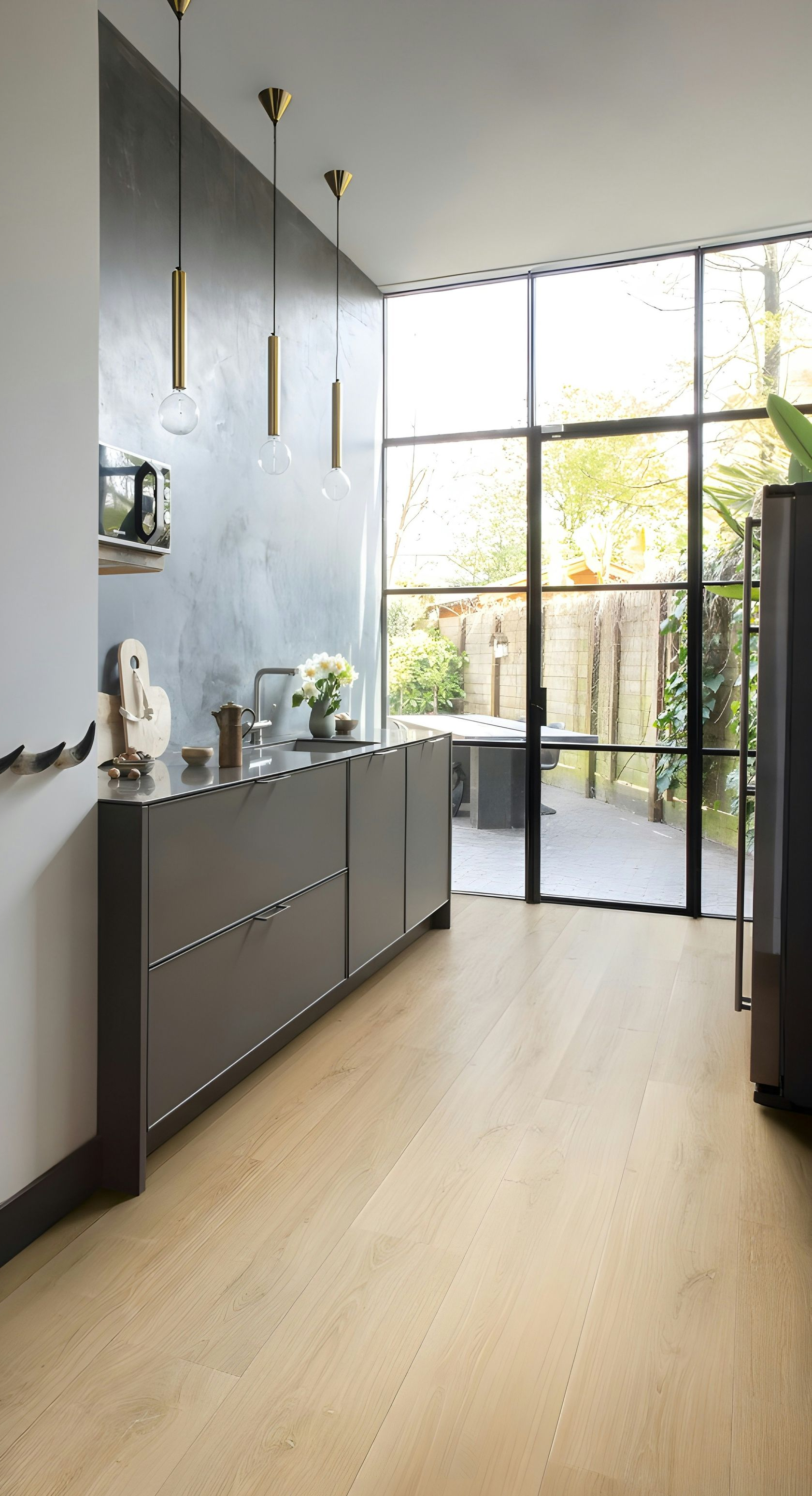 Bright modern kitchen with gray cabinets, light wood floors, and large windows opening to a patio.
