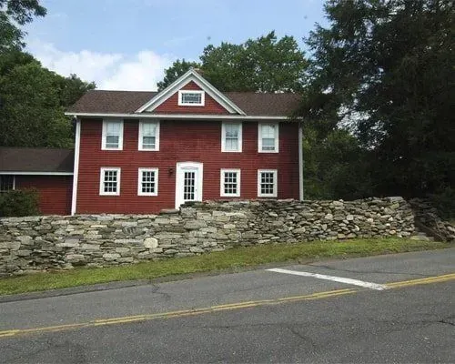 Red clapboard house with white trim behind a stone wall beside a road.