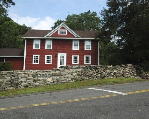 Red two-story house with white trim behind a stone wall beside a road