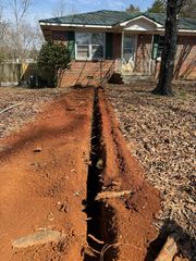 A long trench cut through a yard leads toward a brick house with a porch and trees.