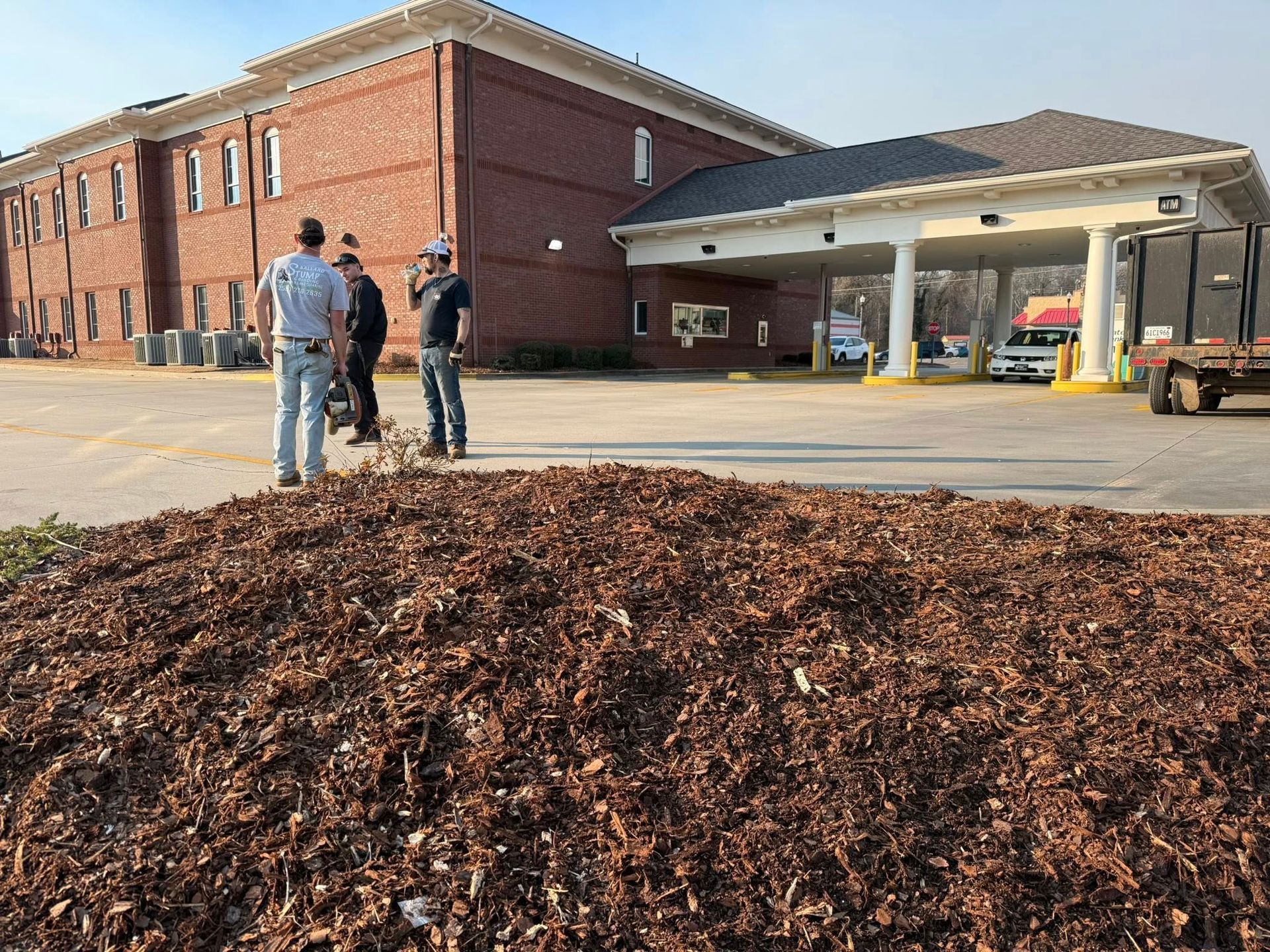 Three people stand by a brick building and driveway beside a large mulch pile on a sunny day