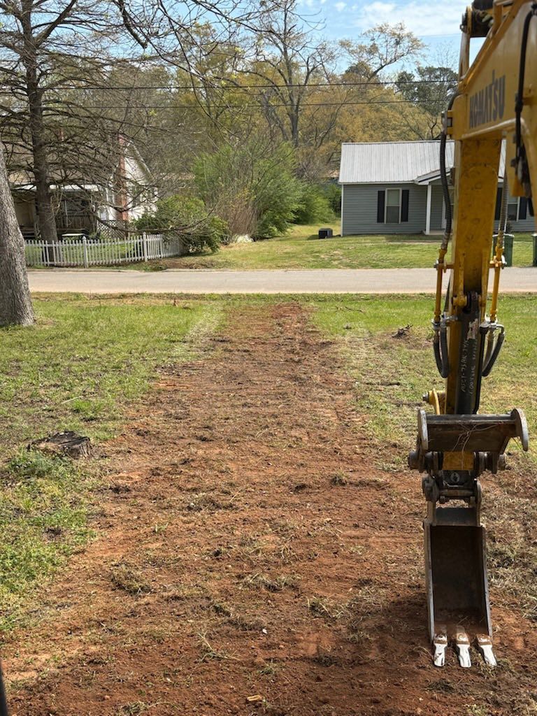 Backhoe bucket beside a freshly cleared dirt path in a grassy yard with trees and a house in the background
