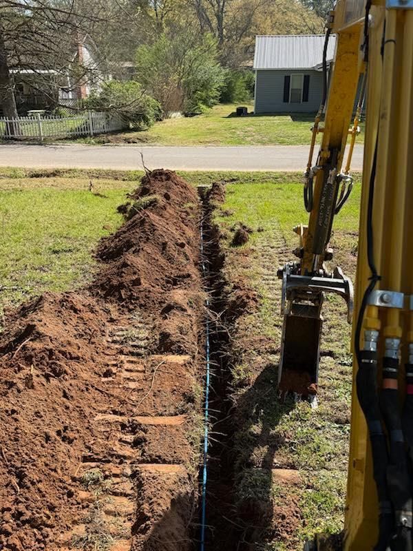 Yellow excavator digging a long trench through a grassy yard beside a suburban street.