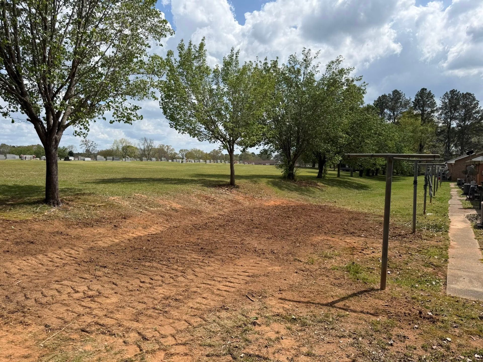 Dirt path beside a grassy field with trees and a cloudy sky, next to a paved walkway.