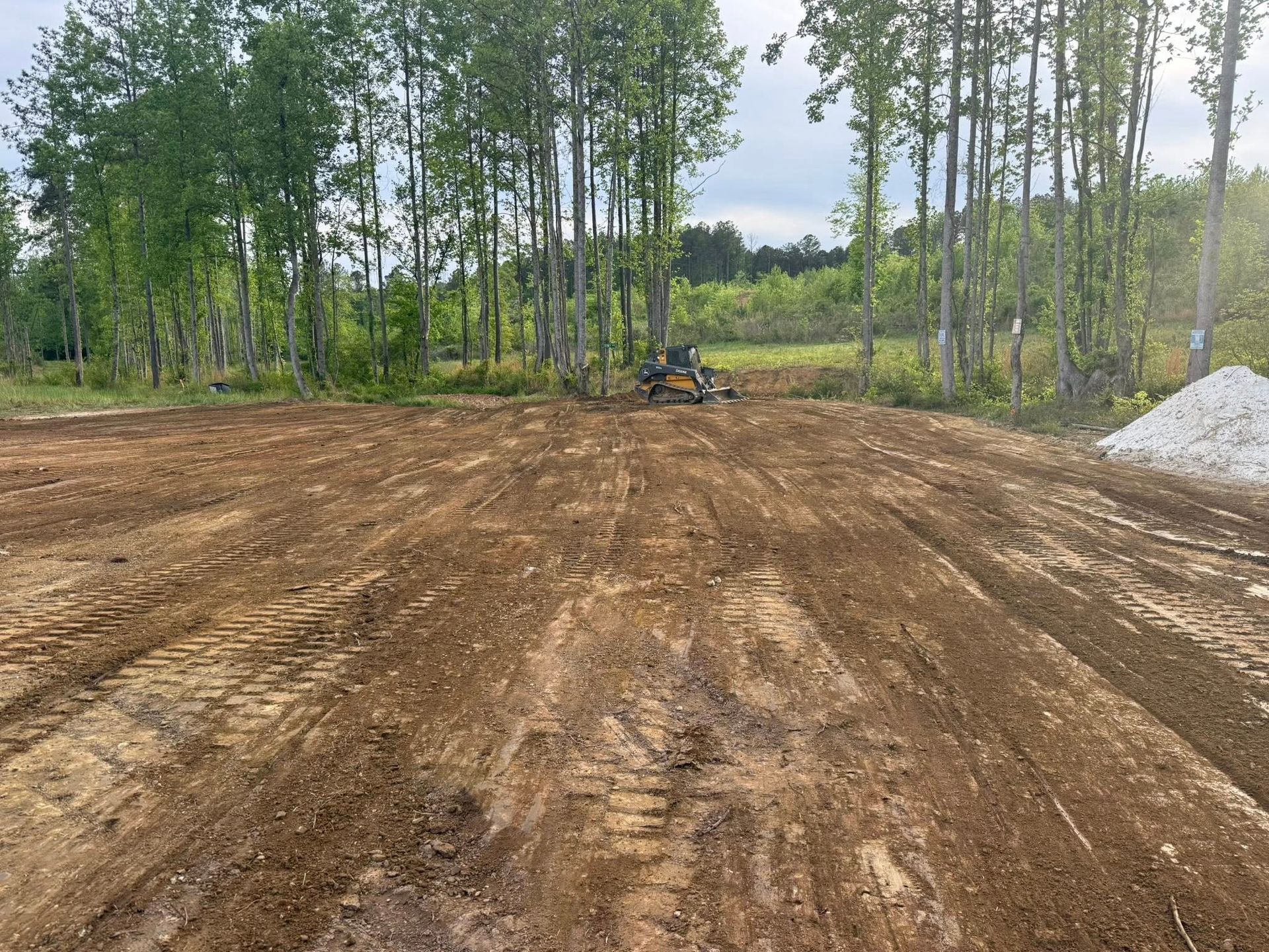 Wide dirt clearing with a person on a small tractor, surrounded by trees and a gravel pile.