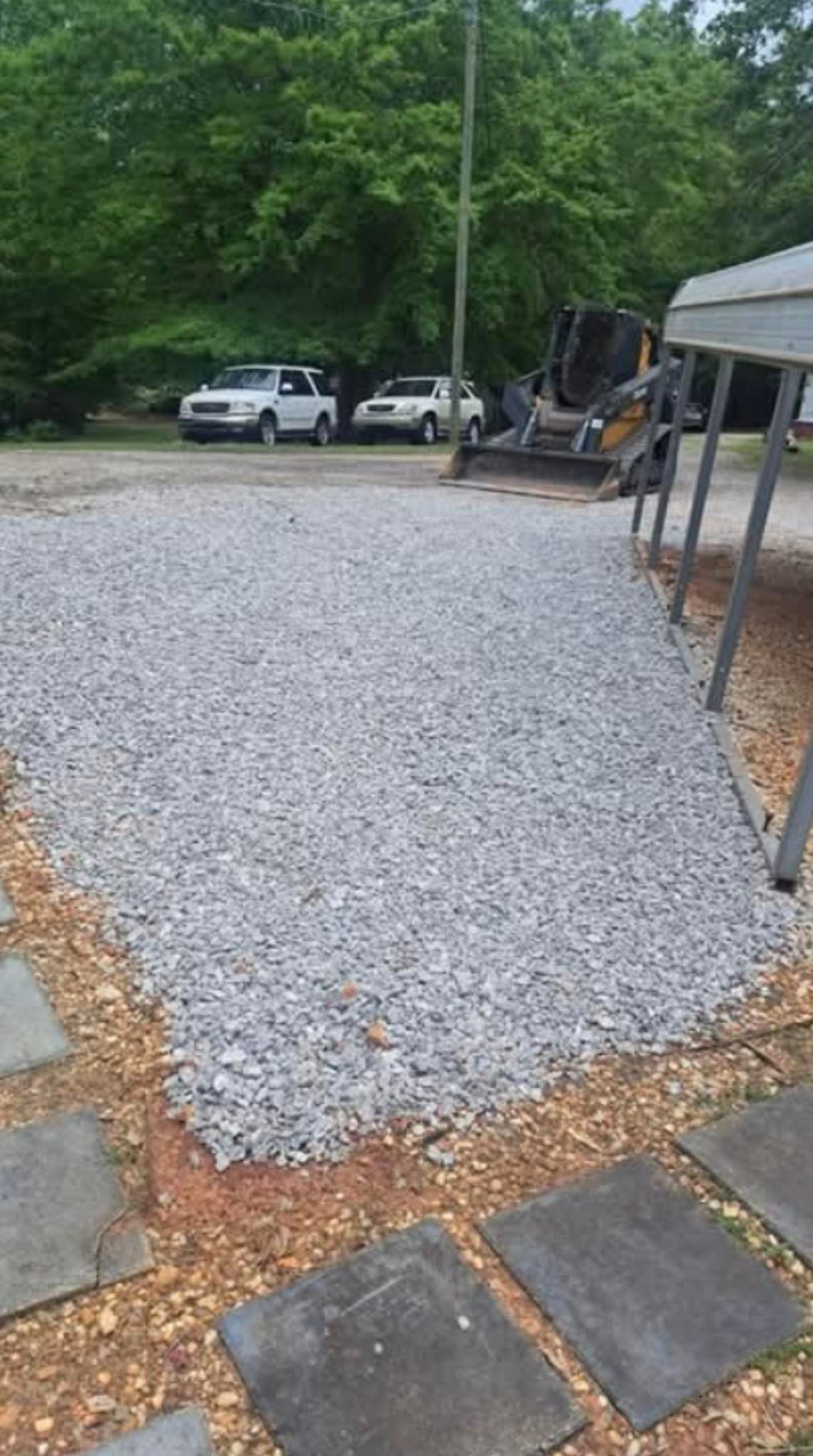 Gravel driveway beside stone pavers, with parked trucks and a bobcat in a wooded yard.