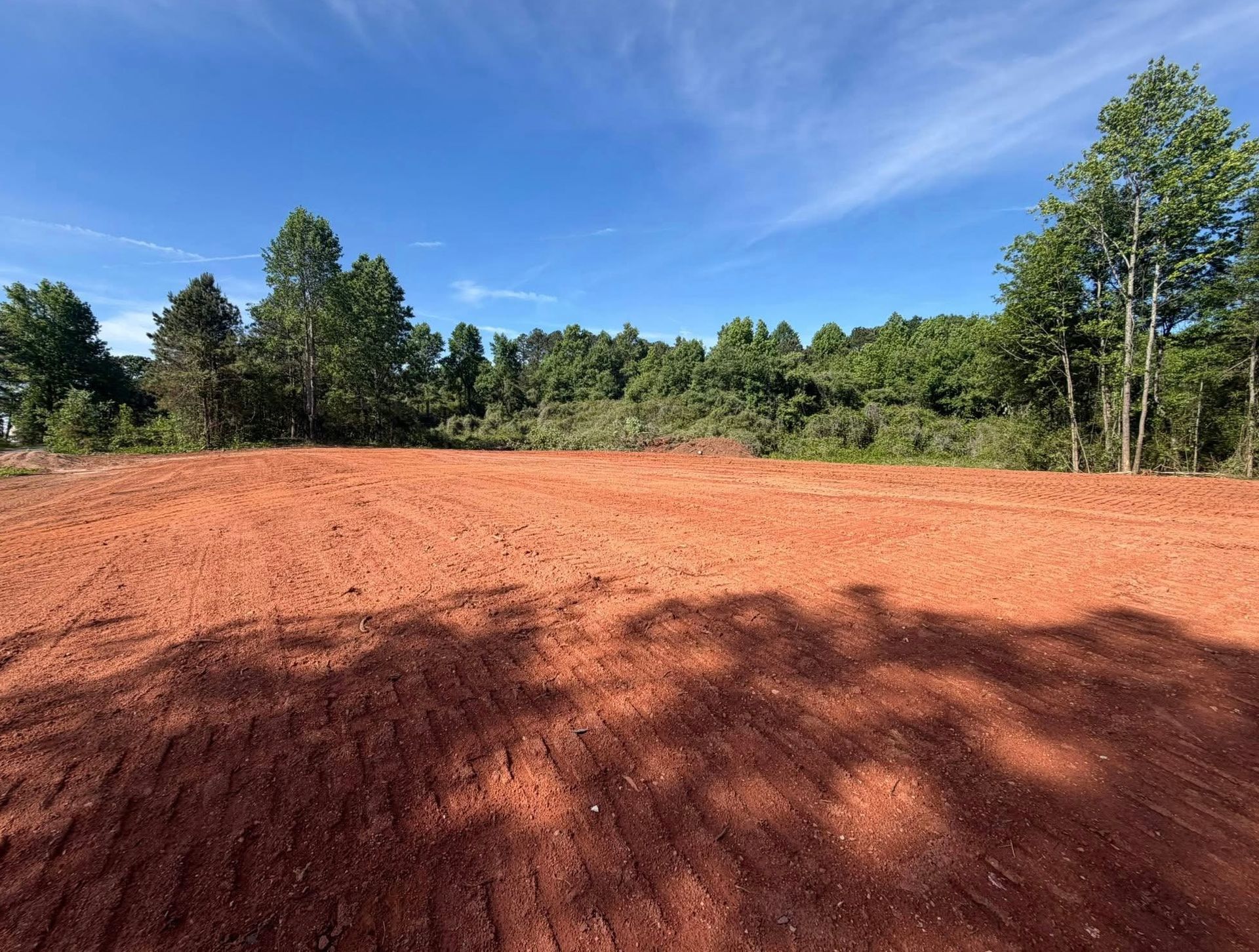 Red dirt clearing bordered by green trees under a bright blue sky with wispy clouds.