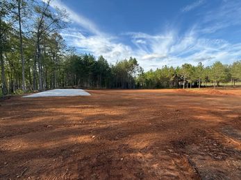 Cleared dirt field bordered by trees under a blue sky with clouds