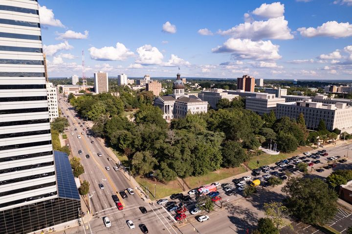 Aerial view of a city street beside a park, with cars, buildings, and a blue sky with clouds.