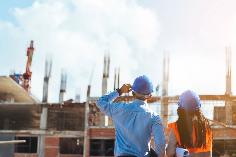 Dos trabajadores de la construcción con cascos examinan una obra en construcción con una grúa bajo un cielo azul.