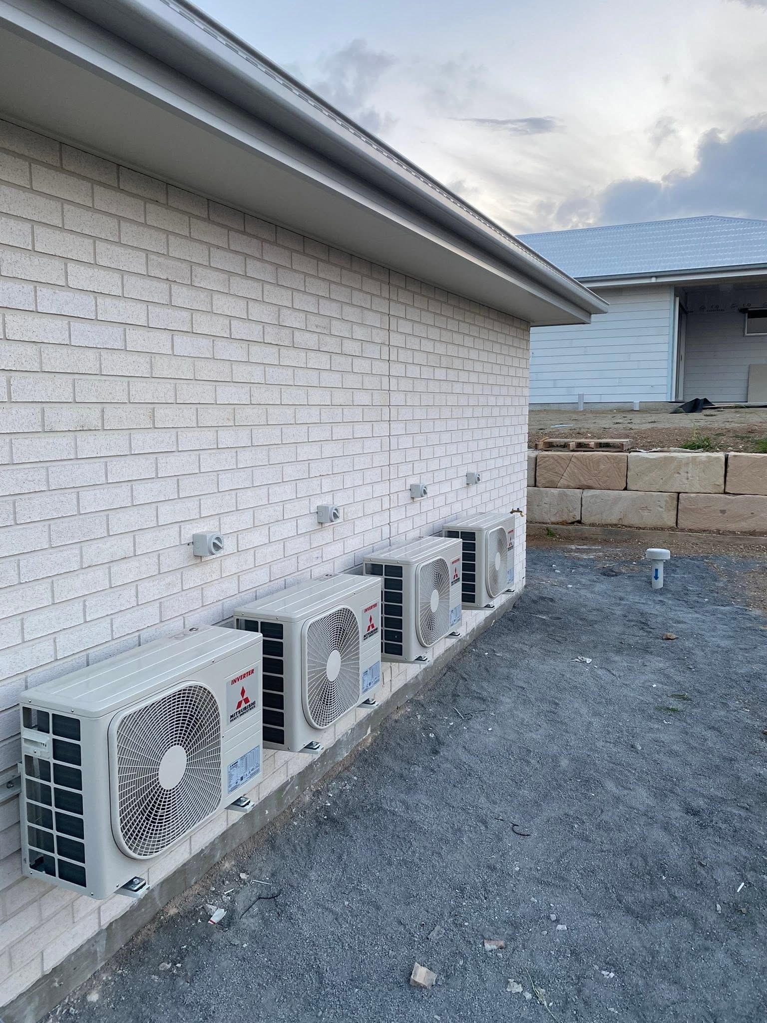 Outdoor wall with four air conditioner units beside a white brick house on gravel.