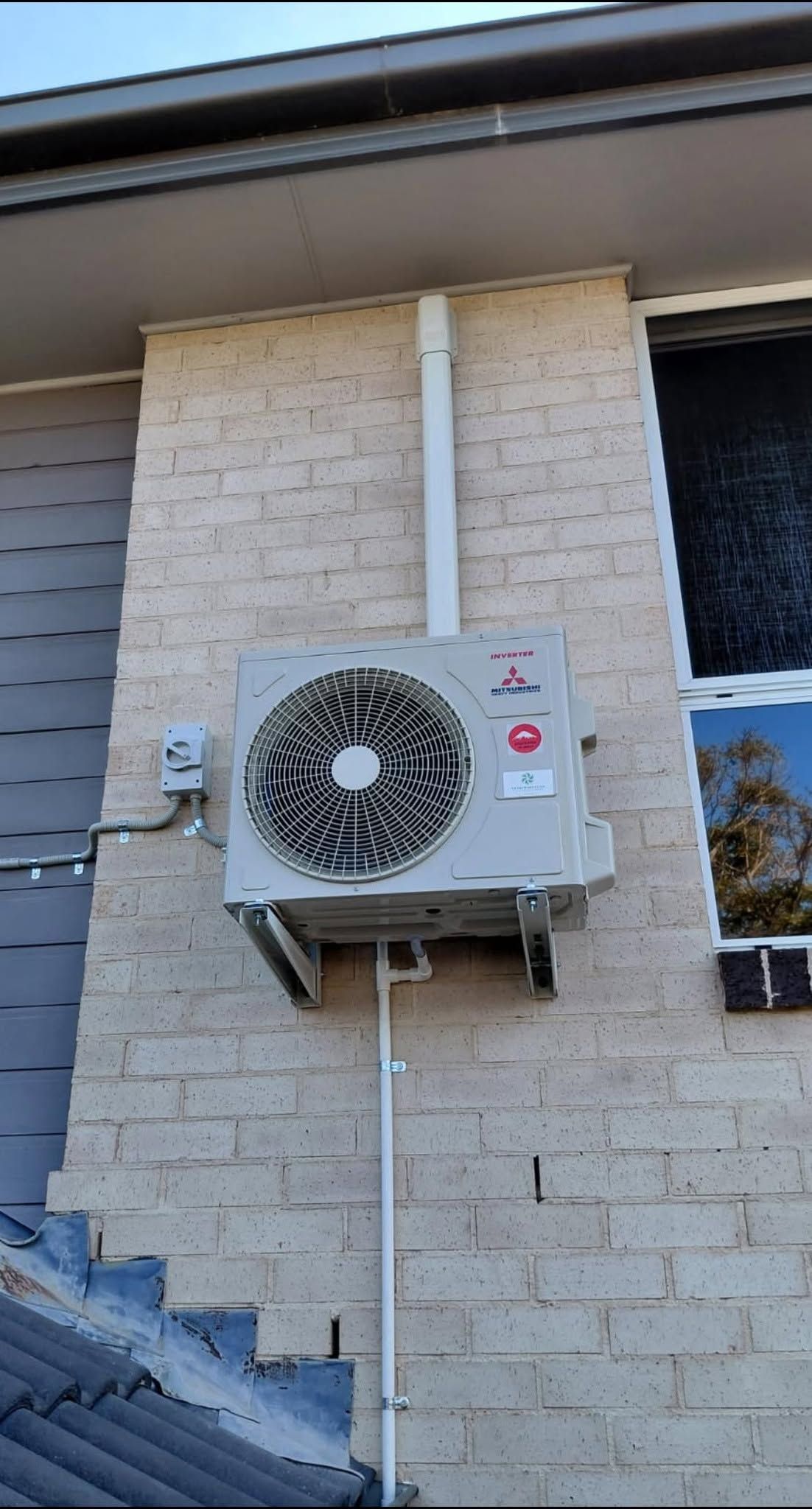 Outdoor air conditioning unit mounted on a beige brick house wall beside a window and roofline