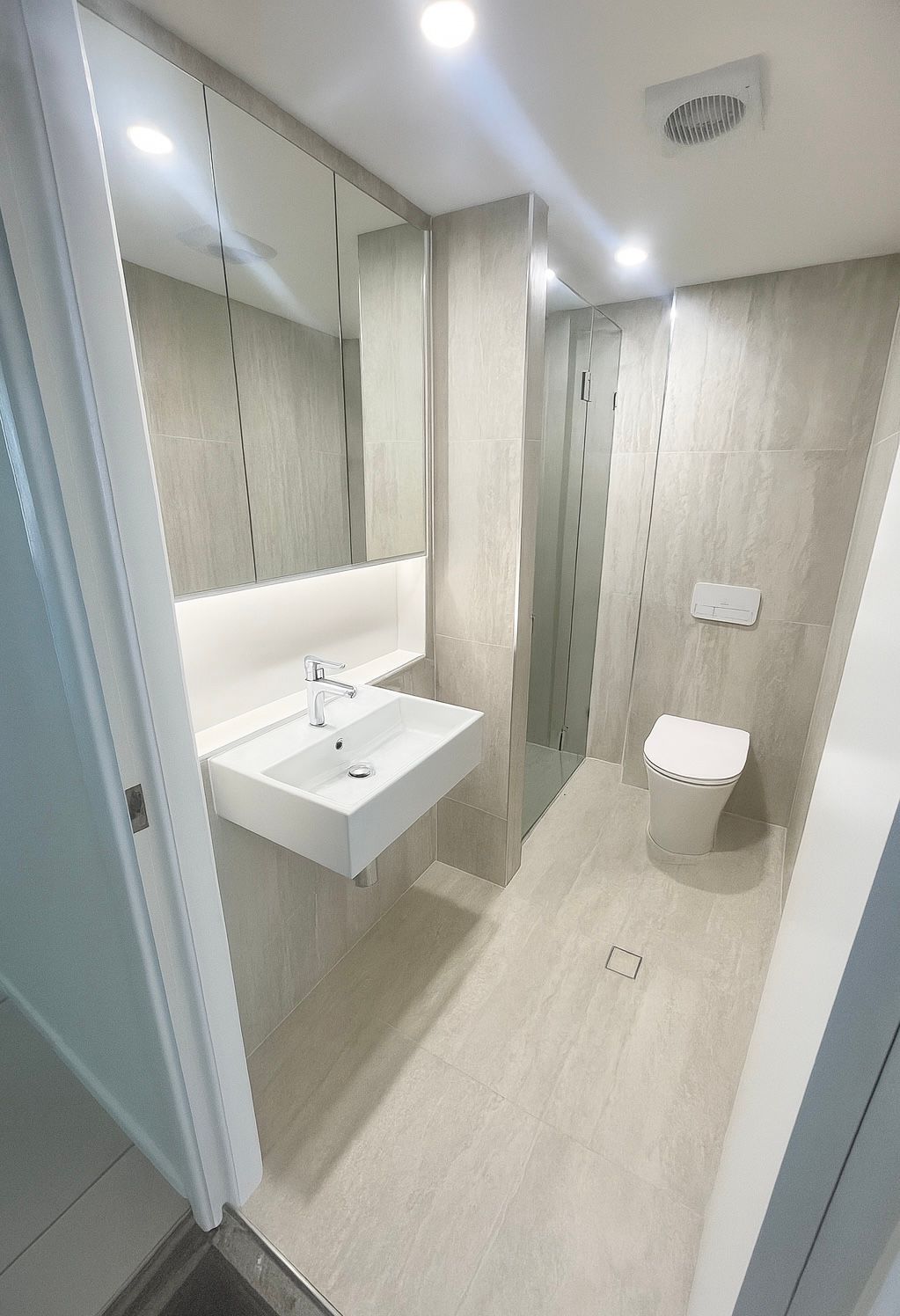 Modern bathroom with a floating sink, toilet, and shower; beige tile, bright lights.