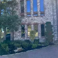 Stone house entrance with arched windows, shrubs, and a driveway in front