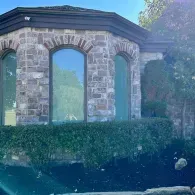 Stone building with arched windows and a dark roof, partly hidden by green hedges and trees.