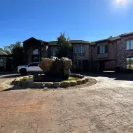Large stone house with a circular driveway and landscaped fountain under a clear blue sky