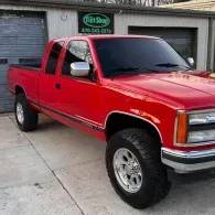 Red lifted pickup truck parked outside a shop, angled front-left view.