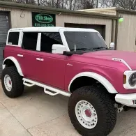 Pink lifted Ford Bronco parked in a driveway in front of a garage