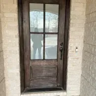 Wooden front door with glass panes set in a beige brick entryway, reflecting a person outside