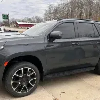 Black SUV parked outdoors on a concrete lot, side view with trees and a red building in the background
