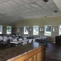 Empty dining hall with rows of tables and chairs, seen from a kitchen counter with windows along the wall