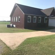 Brick building with white-trimmed windows beside a grassy field and dirt driveway under a blue sky