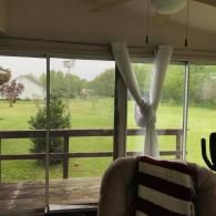 Covered porch with curtains and a chair overlooking a green yard and wooden fence.
