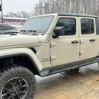 Beige Jeep Gladiator pickup with off-road tires parked on wet pavement in a snowy lot