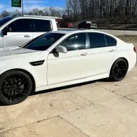 White sedan with black rims parked on a concrete driveway beside a white SUV and trees.