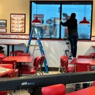 Worker on a ladder cleaning a window inside a restaurant with red chairs and tables.