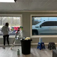 Person cleaning a room with large windows, ladder, trash can, and stools nearby