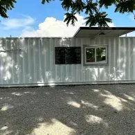 White portable building with a window and sign, shaded by tree branches on a gravel lot