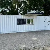 White shipping container café labeled “CHAMPION” with a small window and menu board outdoors.