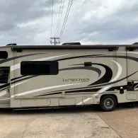 Beige and black luxury motorhome parked on a concrete lot under a cloudy sky