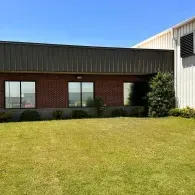 Single-story brick and metal building with windows, a lawn in front, and a tree on the right.