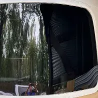 Curved bus window reflecting trees and a person holding up a phone to take a photo
