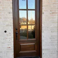 Brown front door with glass panes set in a light brick wall, reflecting trees and sky