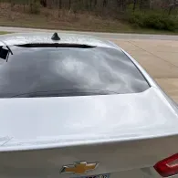 Rear view of a white Chevrolet sedan parked outdoors, with a shattered rear window and visible damage